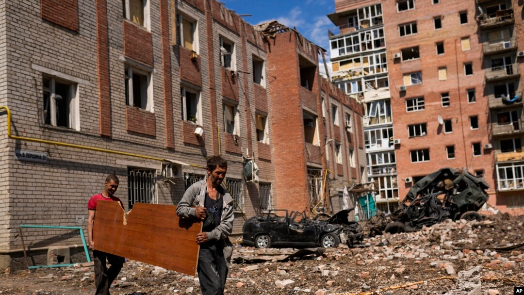 Two men carry a wood panel next to damaged buildings and destroyed cars in a Russian bombing in Bakhmut, eastern Ukraine, May 24, 2022. The town of has been coming under increasing artillery strikes over the last week as Russian forces try to encircle the city of Sieverodonetsk.