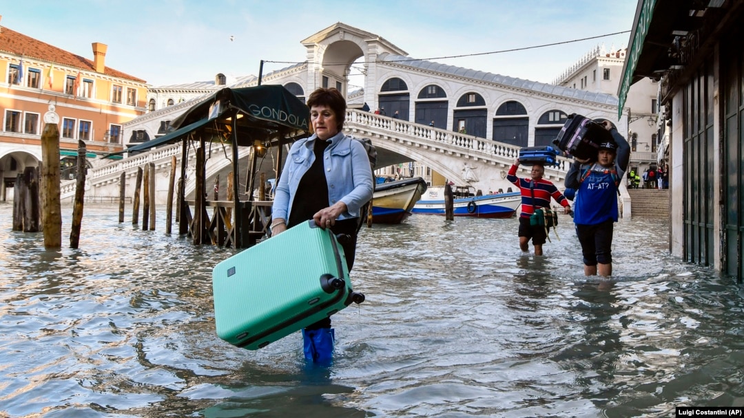 In this Monday, Dec. 23, 2019 file photo, people carry their belongings as they move through water during a high tide of 1.44 meters, near the Rialto Bridge, in Venice, Italy. (AP Photo/Luigi Costantini, file)