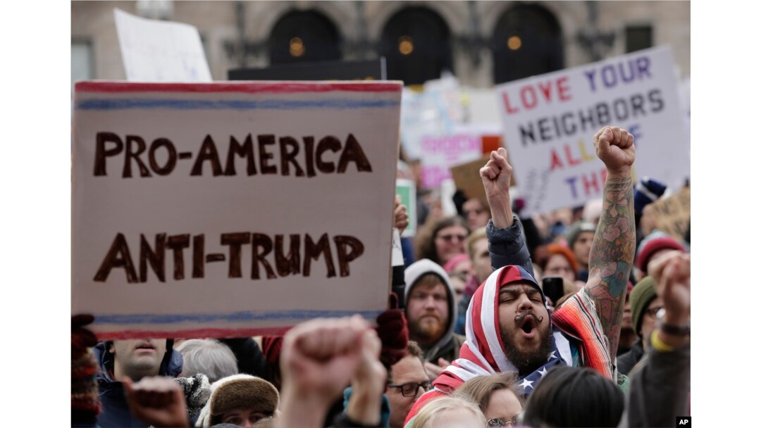 Izzy Berdan, of Boston, center right, wears an American flags as he raises his arm and chants slogans with other demonstrators during a rally against President Donald Trump's order that restricts travel to the US, Jan. 29, 2017, in Boston.